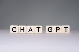 Blocks with letters spell out 'CHAT GPT' on a smooth surface. The blocks are light-colored with black letters, arranged in two groups.