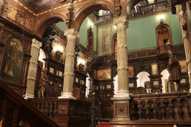 An ornately decorated interior with tall pillars, intricate wood carvings, and classical Renaissance artwork. The space is illuminated by warm lighting, highlighting the opulent architectural details and statues positioned throughout the room.