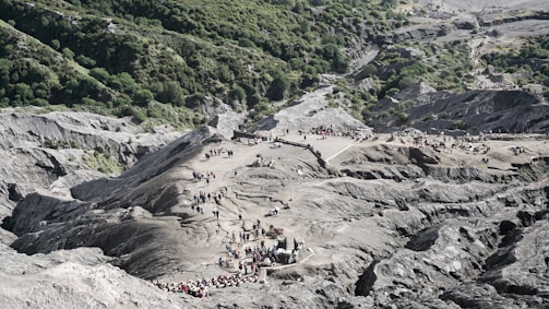 A rugged landscape of volcanic terrain with groups of people walking and gathering in open spaces. The scene is accentuated by lush green forest areas on one side, contrasting with the rocky, gray surface. The presence of smaller peaks and pathways is visible among the larger formations.