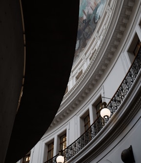 An interior architectural scene featuring a curved, ornate balcony with a wrought-iron railing. Above, intricate molding and a partial view of a mural on the ceiling are visible. Hanging lights illuminate the space, and the contrast between light and shadow adds depth to the scene.