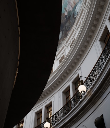 An interior architectural scene featuring a curved, ornate balcony with a wrought-iron railing. Above, intricate molding and a partial view of a mural on the ceiling are visible. Hanging lights illuminate the space, and the contrast between light and shadow adds depth to the scene.