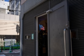 An exterior view of a building with an open door, revealing a neon sign with the letters resembling 'USA' above a depiction of waves or stripes. The street in front has green and white traffic cones and fencing, suggesting construction or maintenance work. Shadows and strong sunlight create a high-contrast scene.