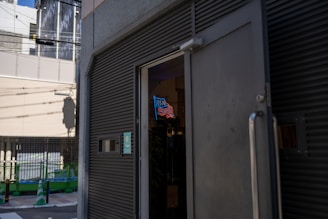 An exterior view of a building with an open door, revealing a neon sign with the letters resembling 'USA' above a depiction of waves or stripes. The street in front has green and white traffic cones and fencing, suggesting construction or maintenance work. Shadows and strong sunlight create a high-contrast scene.