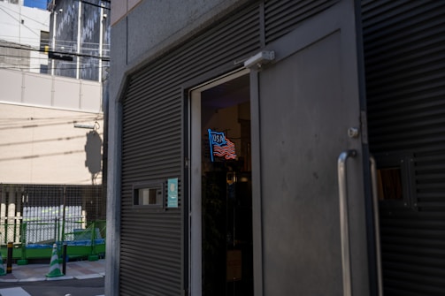 An exterior view of a building with an open door, revealing a neon sign with the letters resembling 'USA' above a depiction of waves or stripes. The street in front has green and white traffic cones and fencing, suggesting construction or maintenance work. Shadows and strong sunlight create a high-contrast scene.