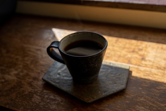 A dark cup of coffee sits on a square coaster placed on a wooden table. Sunlight streams through a window illuminating part of the table and creating a cozy atmosphere.