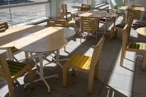 A sunlit indoor seating area with multiple round wooden tables and matching chairs with colorful striped cushions. The room has large windows allowing natural light to create patterns on the tiled floor.