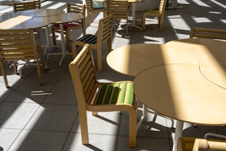 A series of light wooden tables and chairs are arranged on a tiled floor. The chairs have striped cushions with colors like green and blue, and sunlight casts bold shadows across the scene, creating a pattern on the floor.