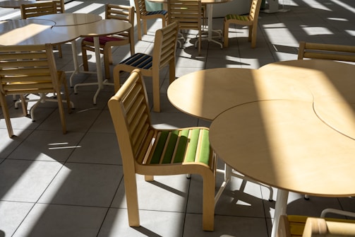 A series of light wooden tables and chairs are arranged on a tiled floor. The chairs have striped cushions with colors like green and blue, and sunlight casts bold shadows across the scene, creating a pattern on the floor.