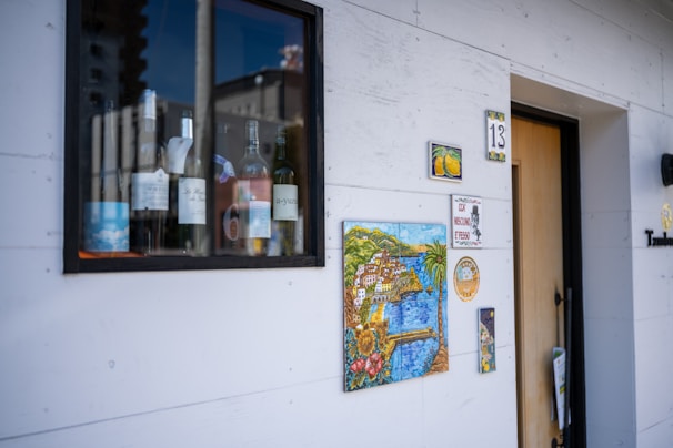 A small window display shows several bottles of wine alongside a mug. Various artistic tiles, including one with a coastal landscape, adorn the white wall next to a doorway numbered 13.