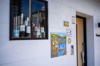 A small window display shows several bottles of wine alongside a mug. Various artistic tiles, including one with a coastal landscape, adorn the white wall next to a doorway numbered 13.