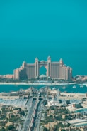 A luxurious hotel with grand architecture featuring towers and an arch design. The waterfront setting includes a bridge leading to the building, with clear blue skies and a tranquil ocean in the background. The surrounding area showcases an urban environment with greenery and a variety of buildings.