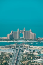 A luxurious hotel with grand architecture featuring towers and an arch design. The waterfront setting includes a bridge leading to the building, with clear blue skies and a tranquil ocean in the background. The surrounding area showcases an urban environment with greenery and a variety of buildings.