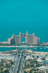 A luxurious hotel with grand architecture featuring towers and an arch design. The waterfront setting includes a bridge leading to the building, with clear blue skies and a tranquil ocean in the background. The surrounding area showcases an urban environment with greenery and a variety of buildings.