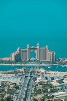 A luxurious hotel with grand architecture featuring towers and an arch design. The waterfront setting includes a bridge leading to the building, with clear blue skies and a tranquil ocean in the background. The surrounding area showcases an urban environment with greenery and a variety of buildings.