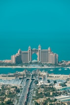 A luxurious hotel with grand architecture featuring towers and an arch design. The waterfront setting includes a bridge leading to the building, with clear blue skies and a tranquil ocean in the background. The surrounding area showcases an urban environment with greenery and a variety of buildings.