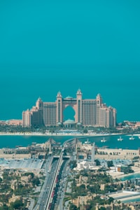 A luxurious hotel with grand architecture featuring towers and an arch design. The waterfront setting includes a bridge leading to the building, with clear blue skies and a tranquil ocean in the background. The surrounding area showcases an urban environment with greenery and a variety of buildings.