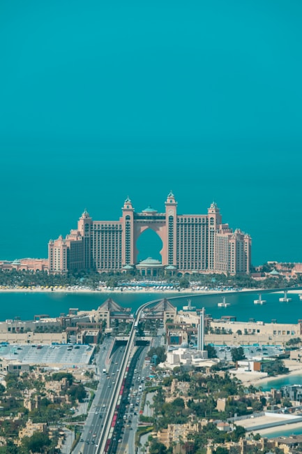 A luxurious hotel with grand architecture featuring towers and an arch design. The waterfront setting includes a bridge leading to the building, with clear blue skies and a tranquil ocean in the background. The surrounding area showcases an urban environment with greenery and a variety of buildings.