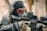 A person dressed in tactical gear, including a skull mask, goggles with orange reflective lenses, and headphones, is aiming a rifle. The gear includes a camouflage uniform and gloves.