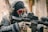 A person dressed in tactical gear, including a skull mask, goggles with orange reflective lenses, and headphones, is aiming a rifle. The gear includes a camouflage uniform and gloves.