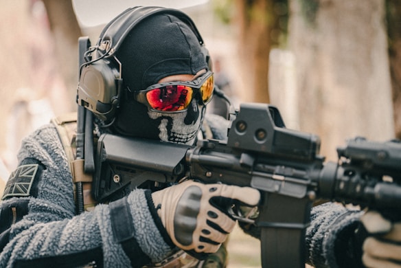 A person dressed in tactical gear, including a skull mask, goggles with orange reflective lenses, and headphones, is aiming a rifle. The gear includes a camouflage uniform and gloves.