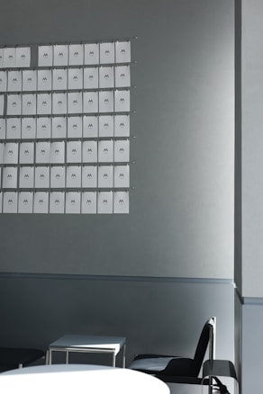 A grid of paper sheets, each featuring the letter 'M', is mounted on a light gray wall. Below the wall, there are minimalistic furnishings, including a white table and a black chair, positioned in a predominantly empty room.