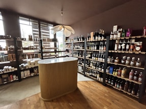A small wine shop or liquor store interior with wooden shelves filled with various bottles of wine and spirits. There is a central, elegantly designed countertop with vertical ridges, and a modern pendant light fixture above it. Natural light streams in through large windows on the left, providing ample illumination. The floor is wooden, adding to the cozy and sophisticated atmosphere.