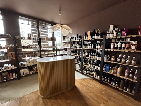 A small wine shop or liquor store interior with wooden shelves filled with various bottles of wine and spirits. There is a central, elegantly designed countertop with vertical ridges, and a modern pendant light fixture above it. Natural light streams in through large windows on the left, providing ample illumination. The floor is wooden, adding to the cozy and sophisticated atmosphere.