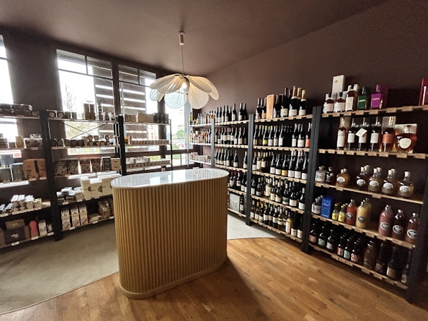 A small wine shop or liquor store interior with wooden shelves filled with various bottles of wine and spirits. There is a central, elegantly designed countertop with vertical ridges, and a modern pendant light fixture above it. Natural light streams in through large windows on the left, providing ample illumination. The floor is wooden, adding to the cozy and sophisticated atmosphere.