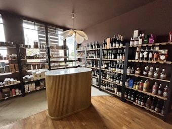 A small wine shop or liquor store interior with wooden shelves filled with various bottles of wine and spirits. There is a central, elegantly designed countertop with vertical ridges, and a modern pendant light fixture above it. Natural light streams in through large windows on the left, providing ample illumination. The floor is wooden, adding to the cozy and sophisticated atmosphere.