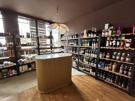 A small wine shop or liquor store interior with wooden shelves filled with various bottles of wine and spirits. There is a central, elegantly designed countertop with vertical ridges, and a modern pendant light fixture above it. Natural light streams in through large windows on the left, providing ample illumination. The floor is wooden, adding to the cozy and sophisticated atmosphere.