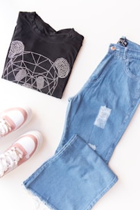 A folded black t-shirt with a rhinestone bear design is placed next to a pair of light blue distressed jeans. Below them, a pair of white sneakers with pink accents are visible. The items are arranged on a white surface, creating a casual outfit layout.