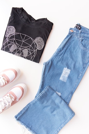 A folded black t-shirt with a rhinestone bear design is placed next to a pair of light blue distressed jeans. Below them, a pair of white sneakers with pink accents are visible. The items are arranged on a white surface, creating a casual outfit layout.