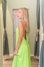 A person with long blonde hair stands sideways in a fitting room, wearing a backless, vibrant green dress. The room is softly lit, with light-colored walls and dark curtains framing the space. There is a door with a subtle design in the background.