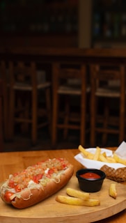 A wooden table holds a plated hot dog topped with diced tomatoes and other vegetables, alongside a basket of golden french fries and a small cup of ketchup. The background shows wooden chairs and a dimly lit bar ambiance.