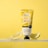 A tube of Farm Stay Lemon Intensive Moisture Foot Cream is positioned upright on a clear surface with splashes of water around it. Two lemon wedges are placed on either side of the tube against a bright yellow background, highlighting the citrus theme.