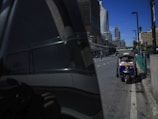 A tuk-tuk is parked on the side of a street in an urban setting. Skyscrapers and modern buildings tower in the background, reflecting in a nearby building's tinted glass. The sky is clear and blue, adding a vibrant contrast to the cityscape.