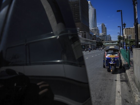 A tuk-tuk is parked on the side of a street in an urban setting. Skyscrapers and modern buildings tower in the background, reflecting in a nearby building's tinted glass. The sky is clear and blue, adding a vibrant contrast to the cityscape.