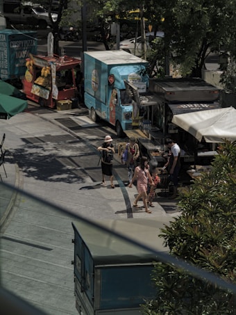 A bustling outdoor food market scene with several food trucks parked along a paved walkway. People can be seen walking and interacting, perhaps deciding on what to eat. The area is shaded by trees, offering some relief from the sun.