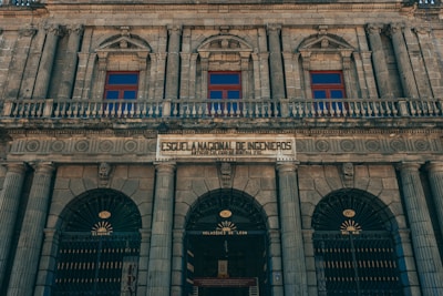 A historic, neoclassical building facade featuring ornate columns and arched windows. The architecture is symmetrical with intricate details, and there is a prominent sign that translates to the 'National School of Engineers.' The structure is constructed from stone, giving it a majestic and timeless appearance.