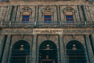 A historic, neoclassical building facade featuring ornate columns and arched windows. The architecture is symmetrical with intricate details, and there is a prominent sign that translates to the 'National School of Engineers.' The structure is constructed from stone, giving it a majestic and timeless appearance.