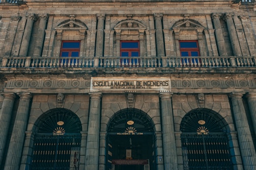 A historic, neoclassical building facade featuring ornate columns and arched windows. The architecture is symmetrical with intricate details, and there is a prominent sign that translates to the 'National School of Engineers.' The structure is constructed from stone, giving it a majestic and timeless appearance.