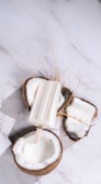 A white popsicle rests across two open coconut halves on a white marble surface. The coconuts are freshly cracked, with white flesh visible inside the rough brown shells and some fibrous strands around them.
