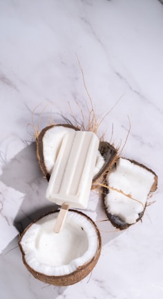 A white popsicle rests across two open coconut halves on a white marble surface. The coconuts are freshly cracked, with white flesh visible inside the rough brown shells and some fibrous strands around them.