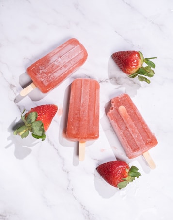 Four strawberry popsicles are arranged on a white marble surface, accompanied by fresh strawberries scattered around them.