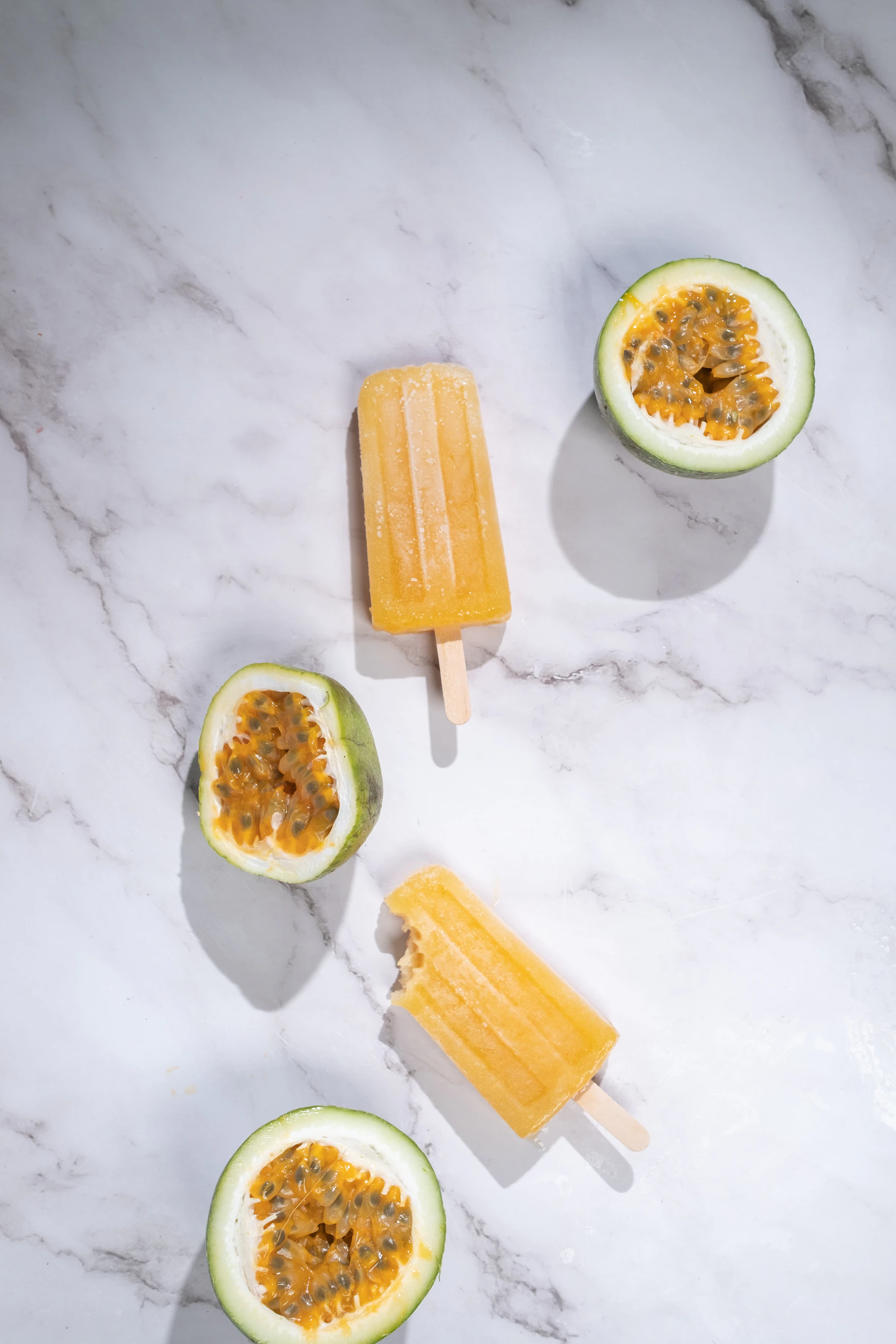 Two pale orange passion fruit pops beside halved passion fruit on white marble