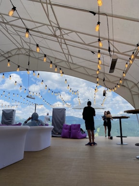 A covered outdoor space featuring string lights under a white tent, with a scenic view of mountains and blue sky. People are seated on white and purple cushioned furniture, while others stand near an umbrella. The overall setting suggests relaxation and social interaction.