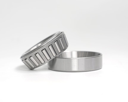 Two metal objects are placed against a white background. One is a cylindrical piece with a smooth finish, lying flat. The other is a tapered roller bearing, positioned at an angle on top of the first object, featuring a series of visible grooves around its circumference.