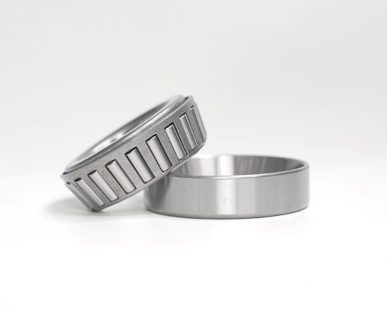 Two metal objects are placed against a white background. One is a cylindrical piece with a smooth finish, lying flat. The other is a tapered roller bearing, positioned at an angle on top of the first object, featuring a series of visible grooves around its circumference.
