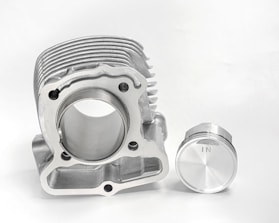 A metallic engine part and a piston are placed against a plain white background. The engine component has a cylindrical bore with multiple ridges on top, highlighting precision engineering. Beside it is a piston with a smooth, shiny surface marked with an 'IN'. Both objects exhibit a clean and polished finish.