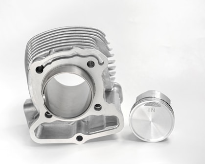 A metallic engine part and a piston are placed against a plain white background. The engine component has a cylindrical bore with multiple ridges on top, highlighting precision engineering. Beside it is a piston with a smooth, shiny surface marked with an 'IN'. Both objects exhibit a clean and polished finish.